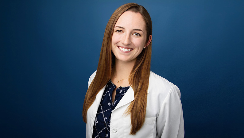Female audiologist wearing white medical coat smiling in front of blue photo background