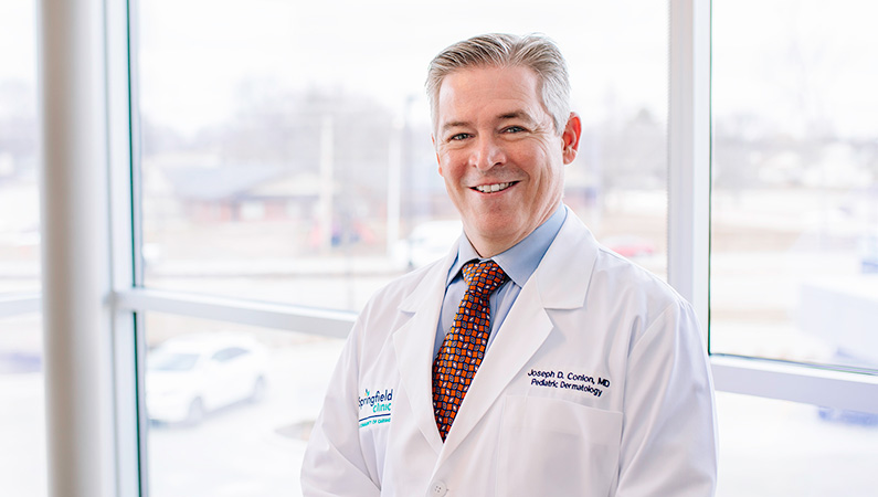 Man wearing a white doctors coat smiling in front of a window