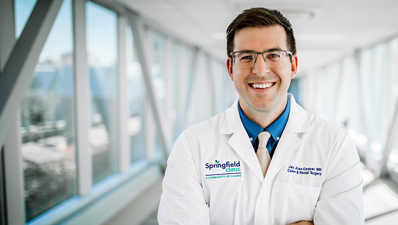 Man wearing a white medical coat smiling in a well-lit walkway.