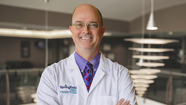 Man wearing a white doctors coat smiling in a well lit lobby.