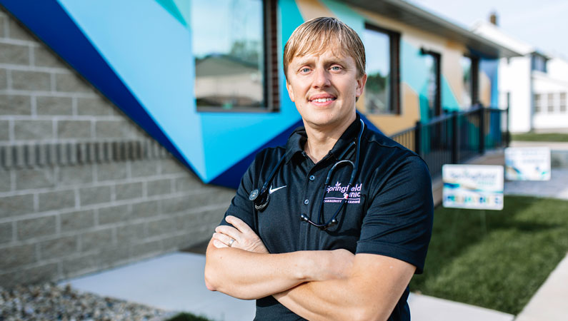Male pediatrics doctor with arms crossed smiling outside in front of building.