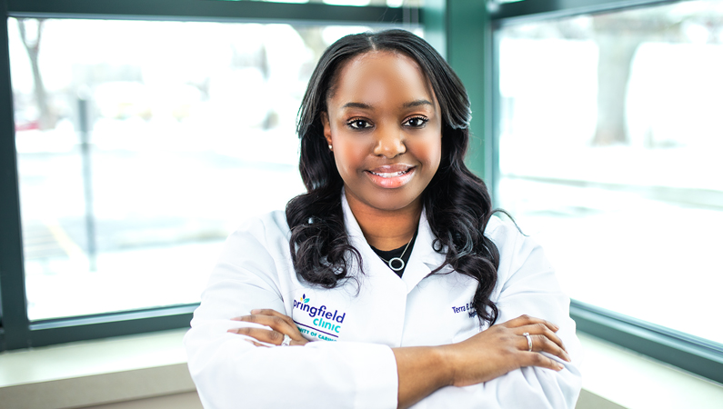 Female with dark hair wearing white medical coat folding arms smiling in front of windows