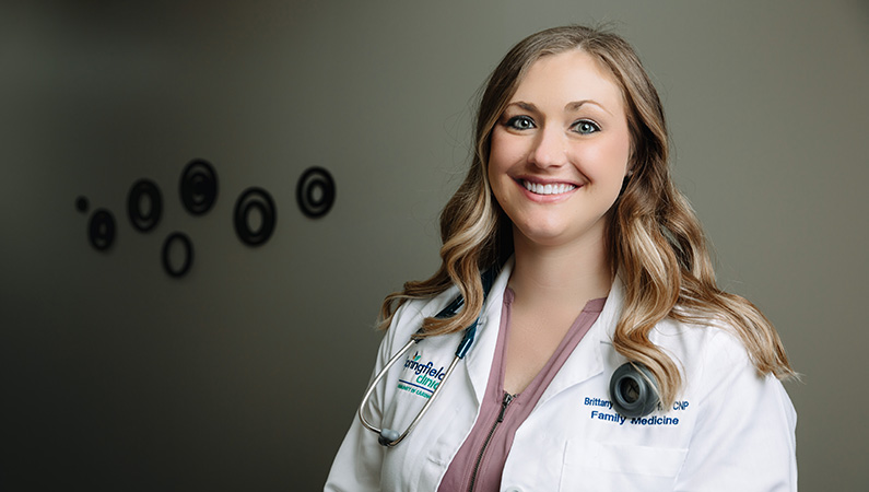 Female nurse practitioner wearing white coat smiling in dark office setting with spotlight on her.