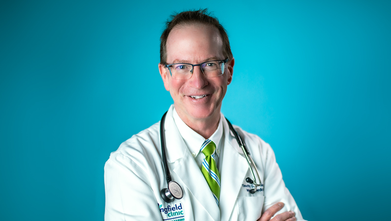 Male wearing glasses and white medical coat with green tie with stethoscope around his neck smiling in front of bright blue background