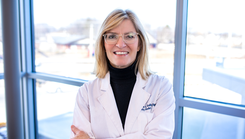 Female with light hair wearing glasses and white medical coat smiling in front of windows