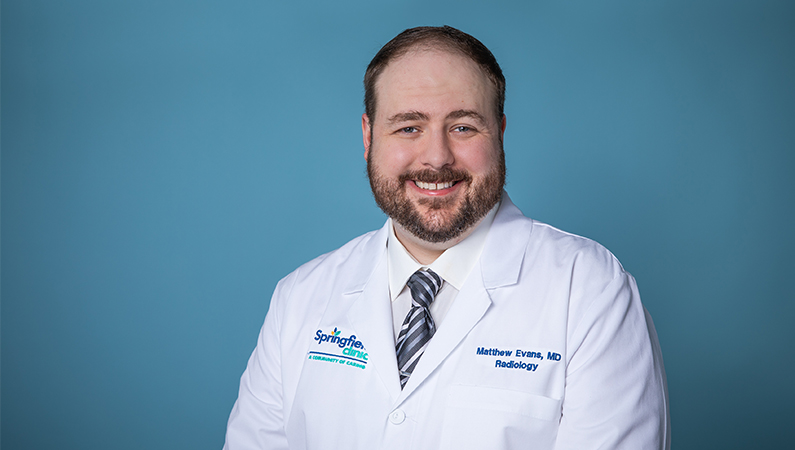 Male radiology doctor in white coat smiling in front of a light blue backdrop.