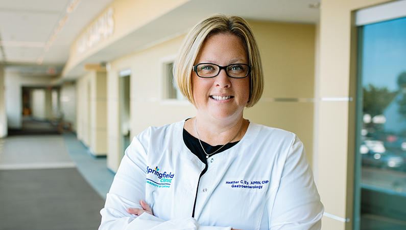 Female with short blonde hair wearing glasses and white medical coat smiling in walkway