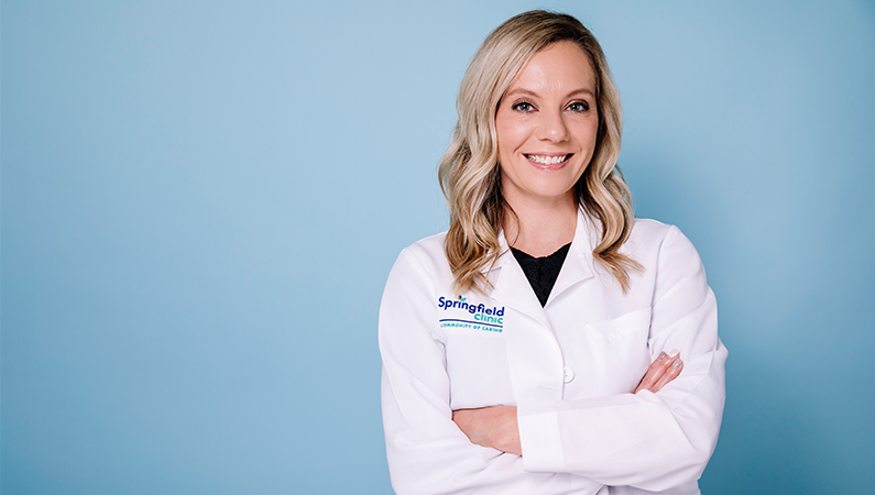 Female plastic surgery nurse practitioner in white coat smiling in front of a blue backdrop.