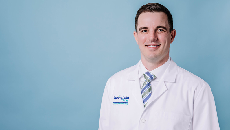 Male nurse practitioner in white coat smiling in front of a blue background.