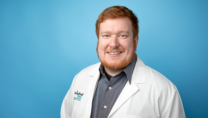 Male nurse practitioner in white medical coat smiling in front of light blue backdrop.