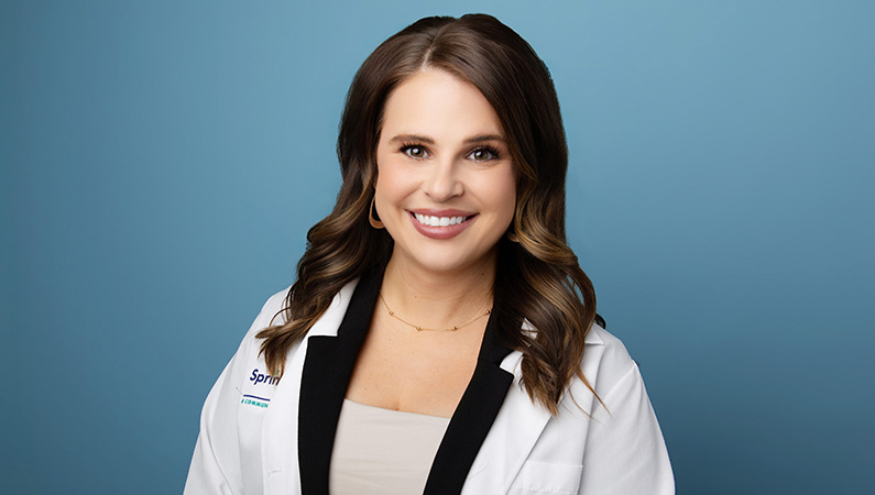 Female nurse practitioner wearing white medical coat smiling in front of blue background