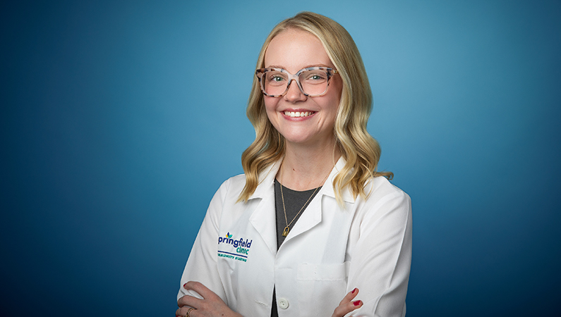 Female nurse practitioner wearing white medical coat smiling in front of blue background