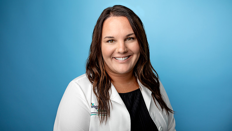 Female nurse in white medical coat smiling in front of light blue photo backdrop.