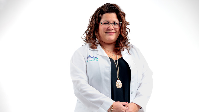 Female nurse practitioner in white medical coat smiling in front of a white photo backdrop.