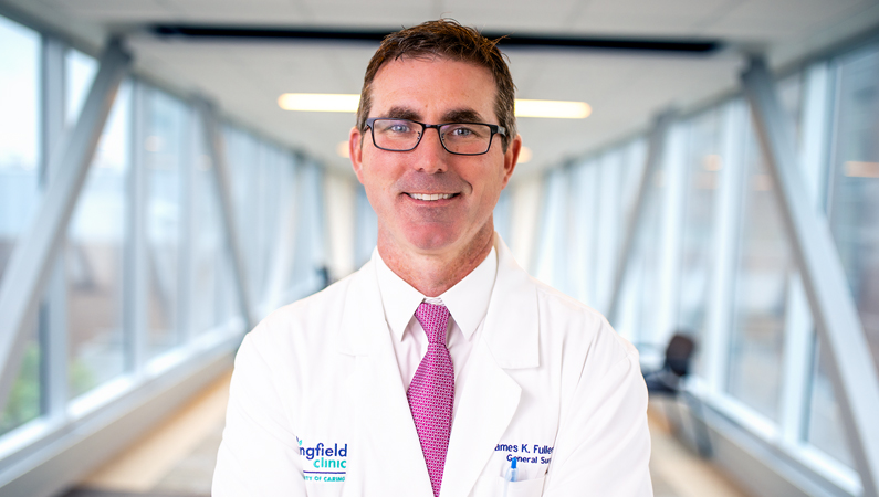 Male with dark hair wearing glasses and white medical coat and red tie smiling in well-lit walkway