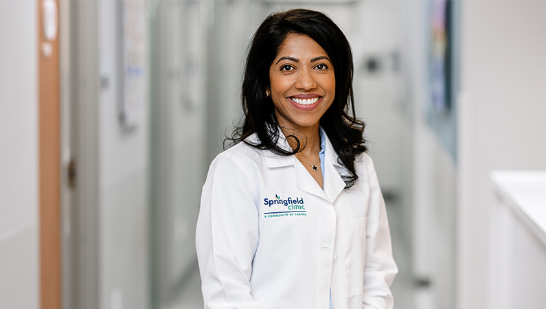 Female doctor in white lab coat smiling in medical facility hallway.