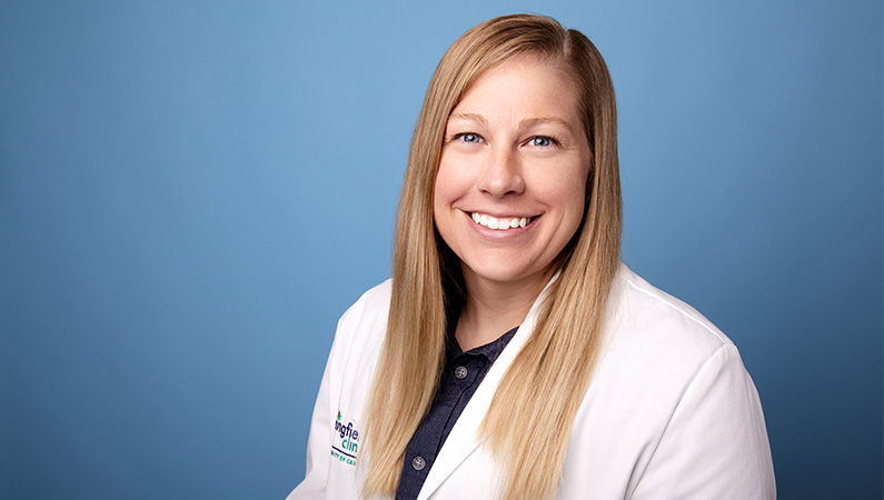 Female nurse practitioner wearing white medical coat smiling in front of blue photo backdrop.