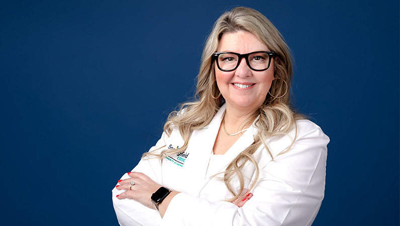 Nurse practitioner in white medical coat smiling in front of blue photo backdrop.
