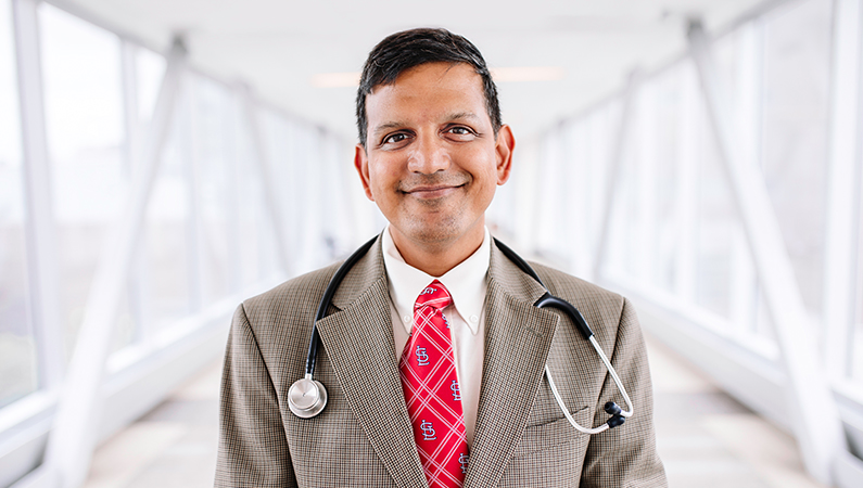 Male wearing suit and tie with stethoscope posing in a well-lit walkway.