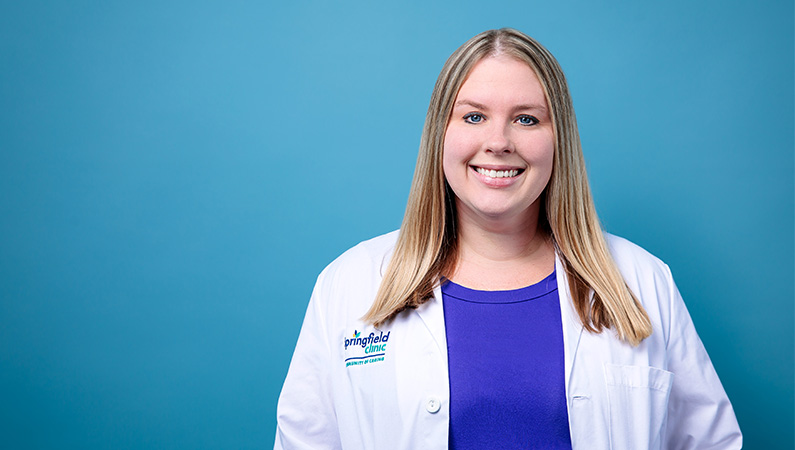 Female nurse practitioner in white medical coat smiling in front of a light blue backdrop.
