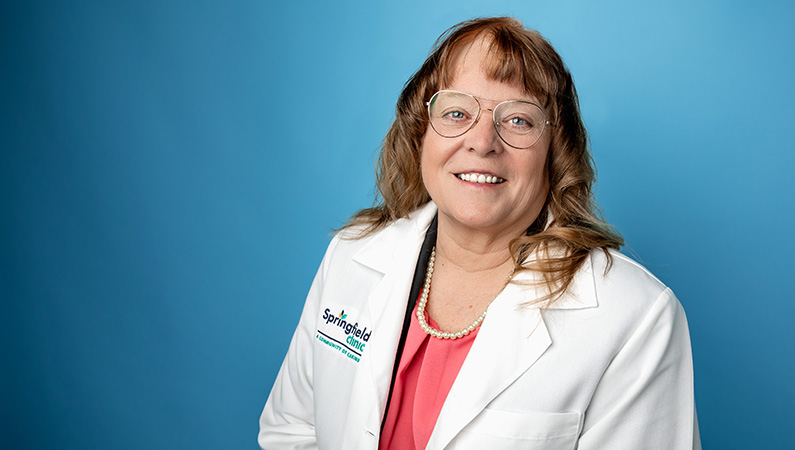 Female cardiology nurse practitioner in white medical coat smiling in front of light blue photo backdrop.