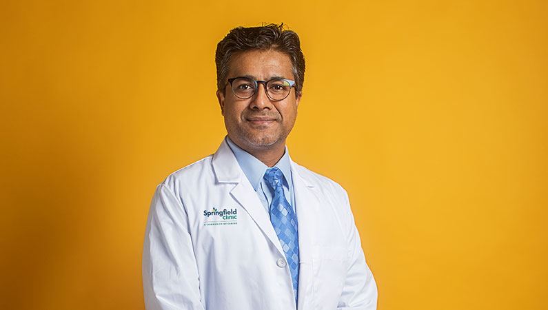 Dark haired male health care provider wearing white coat and glasses posing in front of bright yellow backdrop
