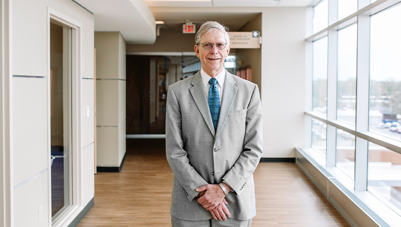 Male internal medicine doctor in grey suit posing in a hallway with glass external windows.