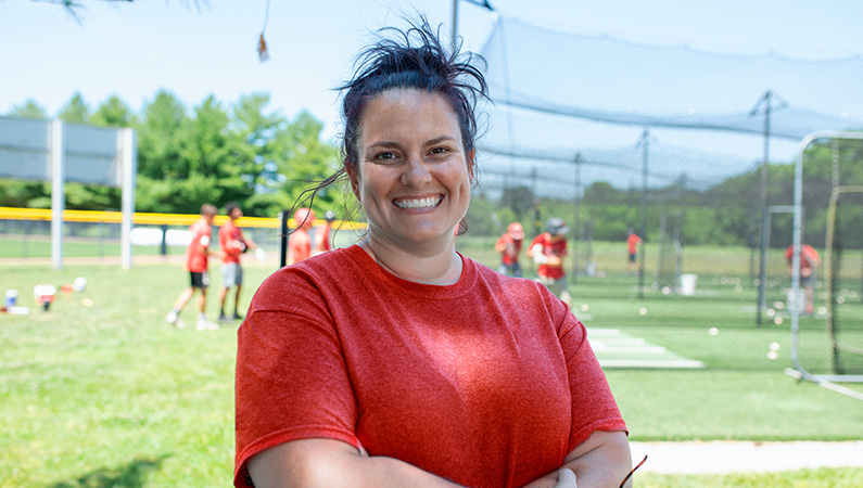 Female with dark hair pulled back wearing red shirt smiling in front of baseball field