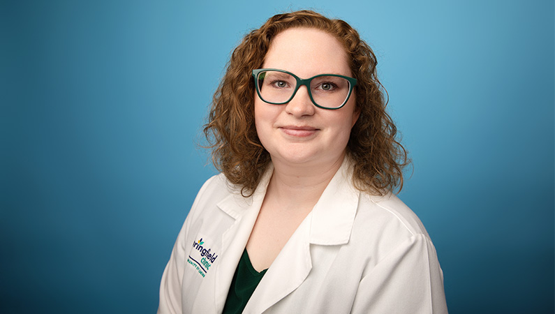 Female nurse wearing white medical coat smiling in front of light blue photo backdrop.