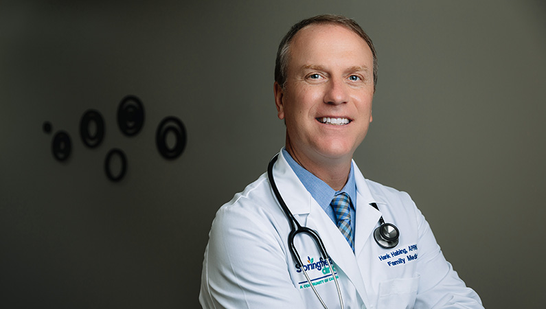 Male nurse practitioner in white coat smiling in a waiting room.
