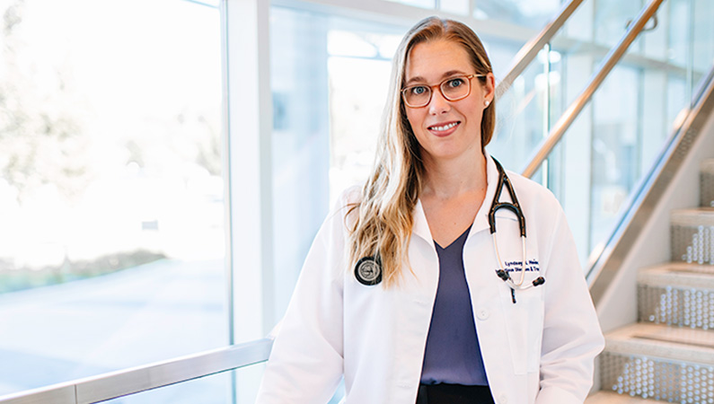Female wearing white medical coat and stethoscope and glasses smiling in front of a staircase