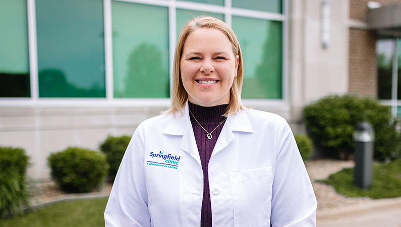 Female with short blonde hair wearing white medical coat smiling in outdoor space in front of medical building