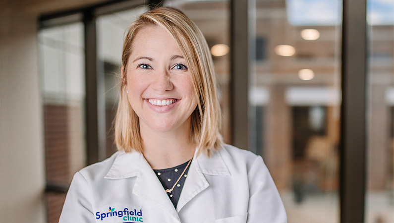 Woman in white doctors coat smiling in front of a window.