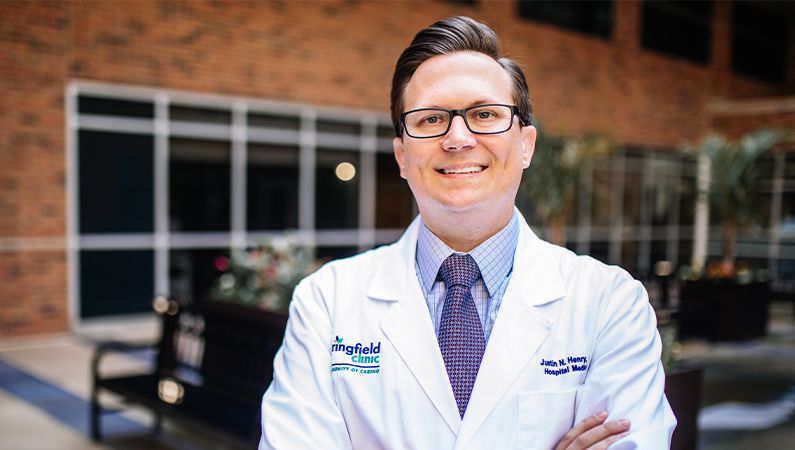 Male wearing glasses and white medical coat smiling in lobby area