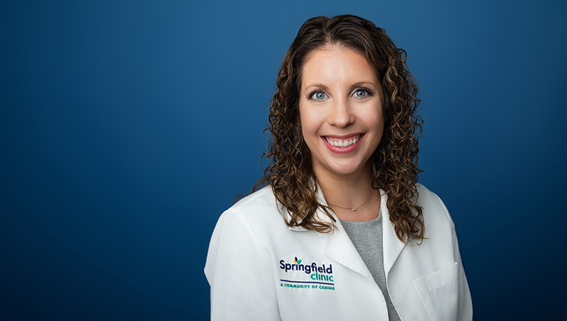 Female physician wearing white medical coat smiling in front of blue photo backdrop.