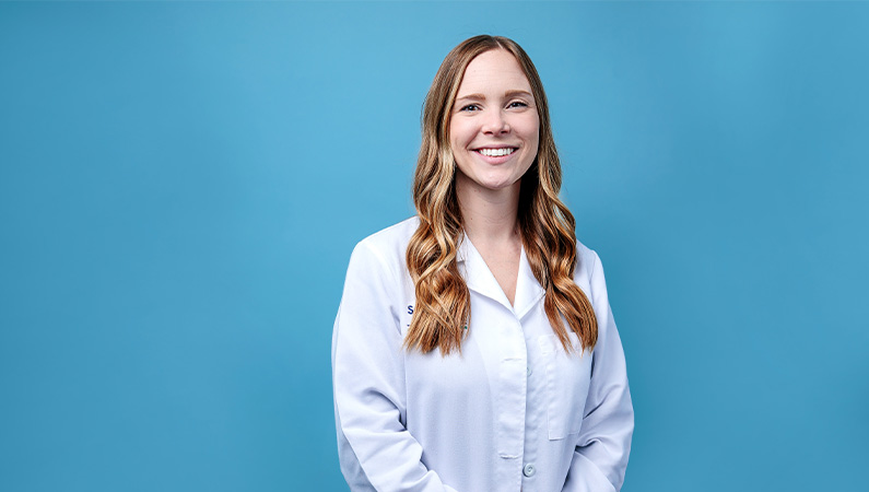 Female nurse practitioner in white medical coat smiling in front of blue photo backdrop.