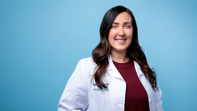 Female neurology nurse practitioner in white lab coat smiling in front of blue photo backdrop.