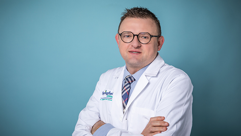Male hospital medicine doctor wearing a white coat posing in front of a light blue backdrop.