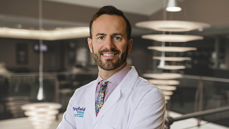 Man wearing a white doctors coat, smiling in a well lit lobby.