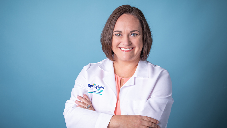 Female nurse practitioner in white coat smiling in front of light blue photo backdrop.