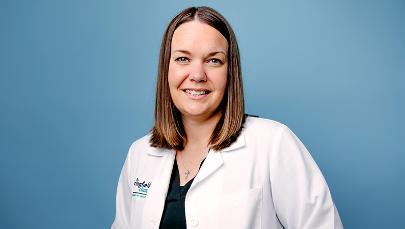 Female nurse practitioner wearing white medical coat smiling in front of light blue photo backdrop.