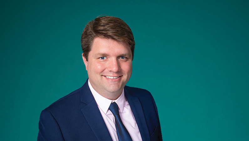 Male with dark hair wearing suit smiling in front of green background