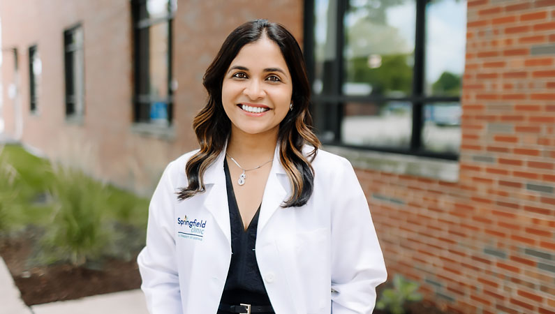 Female doctor in white coat smiling in an outdoor setting in front of a brick building.