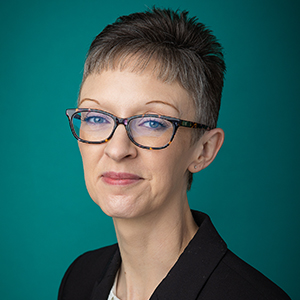 Female nurse practitioner with short hair and glasses smiling in front of green photo backdrop.