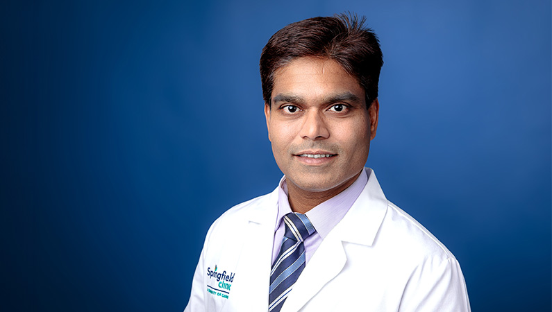 Male doctor wearing white medical coat smiling in front of royal blue photo backdrop.