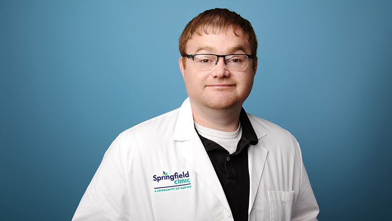 Male nurse practitioner wearing white medical coat posing in front of blue background