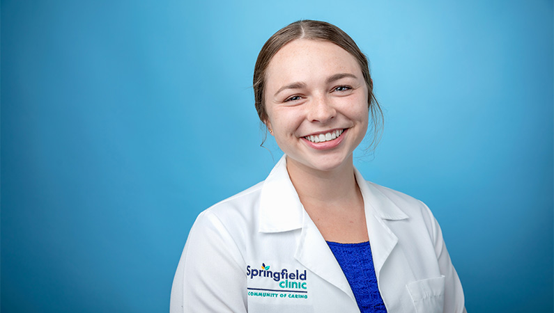 Female physician assistant in white lab coat smiling in front of a light blue photo backdrop.