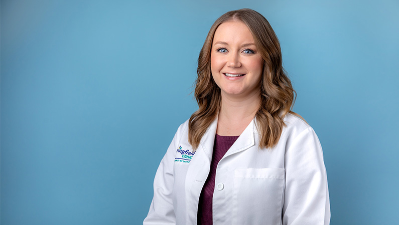 Female nurse practitioner in white coat smiling in front of light blue backdrop.