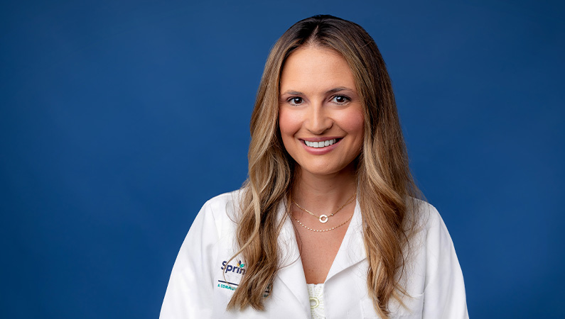 Female doctor wearing white medical coat smiling in front of navy blue background.