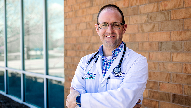 Male wearing glasses and white medical coat smiling posing in outdoor setting